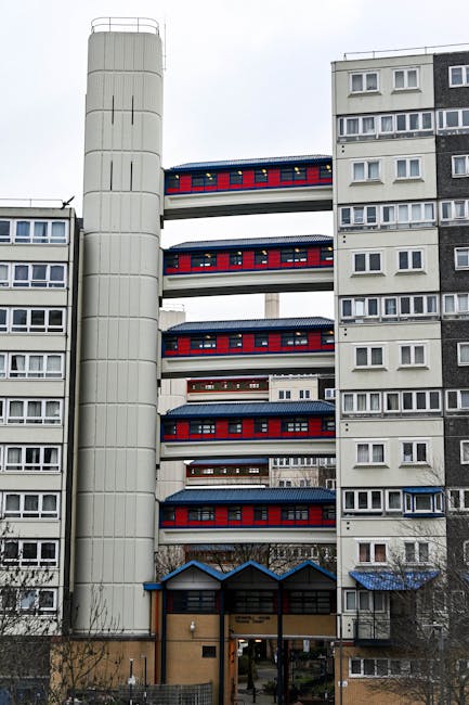 Exterior view of a multi-storey residential building with eight floors, including a ground-level entrance canopy with blue trim. The building has a distinctive external elevator shaft on the left side, structured with vertical white panels, and a skybridge connecting two sections of the building on floors two through six. The façade features a combination of white, grey, and black window frames, with some windows open or partially covered. The building in the image is set against an overcast sky, typical of urban environments, and appears to be part of a housing estate. This setting is relevant for house removals and home relocation services, particularly where access may be restricted by building design and architecture, highlighting the need for specialist moving solutions provided by companies such as Man with Van Kennington.