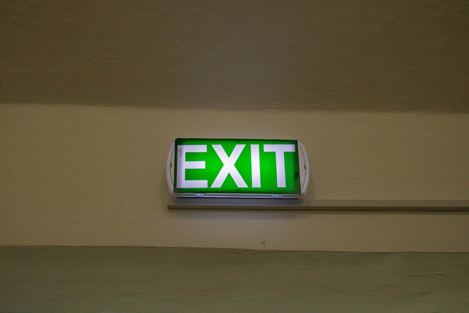 A green illuminated exit sign is mounted on a beige wall inside a property, positioned close to the ceiling. The sign displays the word 'EXIT' in bold white letters. The wall beneath the sign appears smooth, with a horizontal molding or conduit running parallel to the ceiling. The lighting in the area is soft, and there are no other objects or furniture visible in the immediate vicinity. This image could depict a standard indoor corridor or flat interior, relevant to house removals or relocation activities, where clear signage assists in safe navigation during the moving process, as handled by services such as Man with Van Kennington.