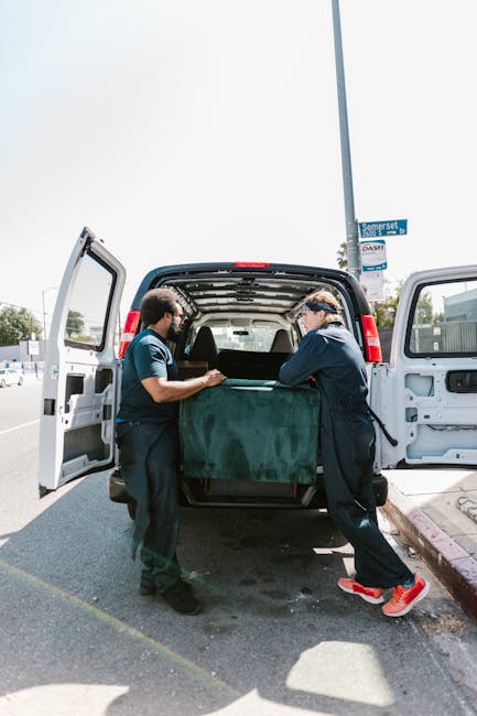 Two movers from Man with Van Kennington are loading a large, dark green upholstered piece of furniture into the open rear of a white panel van, parked on a paved street. The furniture is partially inside the vehicle, with one mover on each side helping to lift and steady it. The inside of the van shows a spacious cargo area, suitable for transporting various household items during a home relocation. The background includes a street sign, trees, and a clear sky, indicating a bright day. The scene captures the careful handling of household goods as part of the furniture transport process, with the movers using proper lifting techniques and working in a coordinated manner. This visual illustrates the logistics involved in packing and moving services offered by [COMPANY_NAME], emphasizing professional furniture transport and loading operations typical of house removals in Kennington.