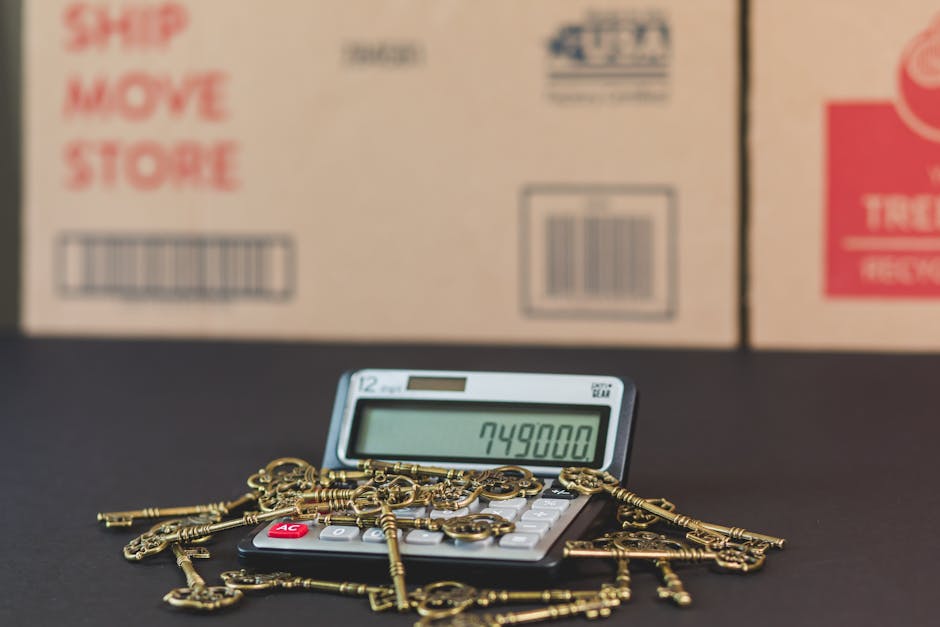 A digital calculator displaying the number 749800 is positioned on a surface with numerous antique brass keys scattered around it, some resting on the calculator's keys. Behind the calculator, there are cardboard boxes with printed labels and barcodes, indicating their use for moving or packing purposes. The setting appears to be a staging area for a home relocation, with the boxes likely containing household items or furniture to be moved. The brass keys, which are of various sizes and shapes, suggest the presence of locks or furniture, and they are loosely arranged, some overlapping each other. The background is out of focus, emphasizing the calculator and keys in the foreground. This scene illustrates the process of moving and storage, where packing materials and key objects are prepared for transport, in association with the professional packing and loading services offered by Man with Van Kennington, particularly for house removals and furniture transport.