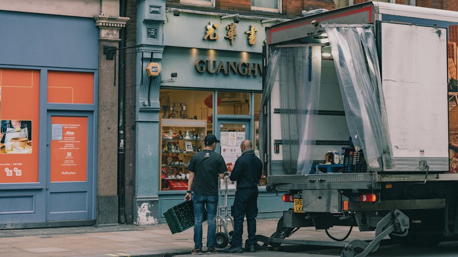 Two movers from Man with Van Kennington are loading a large, dark green upholstered piece of furniture into the open rear of a white panel van, parked on a paved street. The furniture is partially inside the vehicle, with one mover on each side helping to lift and steady it. The inside of the van shows a spacious cargo area, suitable for transporting various household items during a home relocation. The background includes a street sign, trees, and a clear sky, indicating a bright day. The scene captures the careful handling of household goods as part of the furniture transport process, with the movers using proper lifting techniques and working in a coordinated manner. This visual illustrates the logistics involved in packing and moving services offered by [COMPANY_NAME], emphasizing professional furniture transport and loading operations typical of house removals in Kennington.