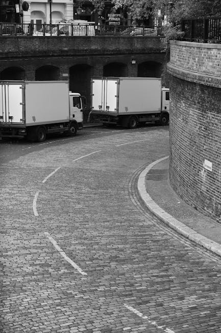 A black and white photograph depicting a curved cobblestone driveway in an urban setting, with two white box trucks parked side by side along the edge of the street. Both trucks have closed cargo door