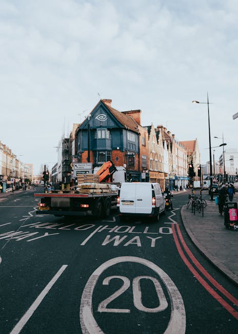 A white van and a flatbed truck parked on a narrow urban street during a home relocation process, with the flatbed loaded with packed cardboard boxes, wooden furniture, and wrapped items. The truck is