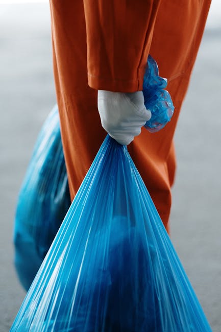A waste collection worker wearing a fluorescent yellow and red uniform is standing behind the open rear loading area of a large red waste collection truck. The worker is handling trash bags, with some