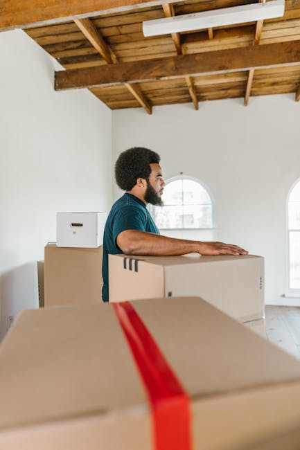 A man with dark skin, curly hair, and a beard is standing inside a room with a white wall, large arched windows, and an exposed wooden ceiling structure. He is wearing a dark blue t-shirt and appears to be in the process of a home relocation, standing behind several brown cardboard boxes of various sizes, some topped with white boxes, which are stacked on top of each other. The boxes are likely sealed with packing tape, indicating they are prepared for moving. The man has his arms resting on one of the boxes, possibly preparing to lift or position them during the packing and moving process. In the background, there is a white doorframe, and the natural light coming through the windows illuminates the space. This scene is typical of the loading phase in furniture transport and packing services offered by [COMPANY_NAME], such as Man with Van Kennington, emphasizing the careful handling and organization involved in the house removals process.