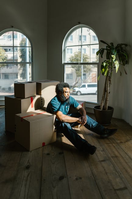 A man with curly hair, wearing a blue t-shirt and dark trousers, is sitting on a wooden floor inside a room with large arched windows that let in natural light. He is surrounded by several cardboard moving boxes, some stacked nearby and others placed on the floor. The boxes are taped and appear to be packed for a home relocation. The room features a tall potted plant with broad green leaves positioned beside the man. Outside the windows, a street with parked cars and residential buildings can be seen. This scene depicts the process of packing and preparing for a moving or house removal, with the man representing a professional mover or someone involved in furniture transport and relocation logistics. The setting suggests a home environment undergoing a packing and loading process as part of the moving services offered by Man with Van Kennington, aligned with tips for last-minute movers and avoiding common mistakes during house moves.