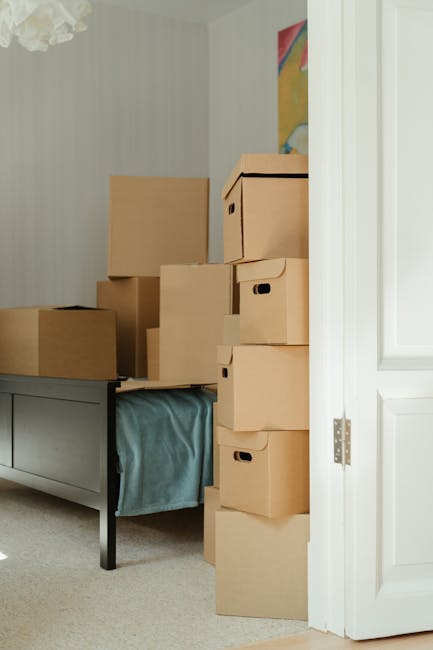 Inside a room during a home relocation, several cardboard boxes of various sizes are stacked on top of a grey piece of furniture with a light blue cloth hanging over the side. The boxes are neatly arranged, some placed directly on the furniture surface and others stacked against a white wall. The room has a patterned wallpaper, partially visible, and a colorful abstract painting hanging on the wall behind the boxes. Natural light illuminates the scene, highlighting the neutral tones of the boxes and furniture. This scene depicts the packing stage of a house move, with the boxes prepared for transportation, likely being handled by a professional moving service such as Man with Van Kennington, specializing in house removals and furniture transport in the Kennington area.