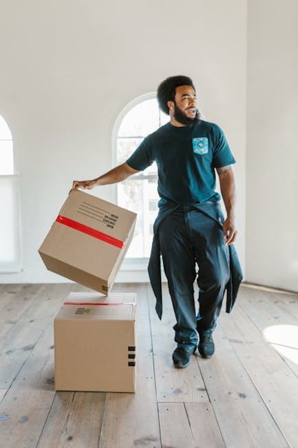 An African man with a beard and natural afro hairstyle is standing inside a bright, spacious room with white walls and large arched windows, during a home relocation or furniture transport process. He is wearing a dark t-shirt with a blue logo on the chest, dark trousers, and has a jacket tied around his waist. The man is holding a cardboard box with red tape, which appears to contain household items or belongings, and is in the act of placing or lifting it onto another box on the wooden floor. The environment is well-lit by natural daylight streaming through the windows, emphasizing the clean, uncluttered interior space. Behind him, the room lacks furniture, suggesting it is either in the process of being packed or unpacked during a house move. This scene is associated with professional removals, packing, and moving services such as those offered by Man with Van Kennington, which specialise in house relocations in the Kennington area.