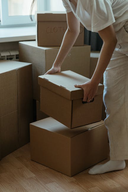 Inside a room during a home relocation, several cardboard boxes of various sizes are stacked on top of a grey piece of furniture with a light blue cloth hanging over the side. The boxes are neatly arranged, some placed directly on the furniture surface and others stacked against a white wall. The room has a patterned wallpaper, partially visible, and a colorful abstract painting hanging on the wall behind the boxes. Natural light illuminates the scene, highlighting the neutral tones of the boxes and furniture. This scene depicts the packing stage of a house move, with the boxes prepared for transportation, likely being handled by a professional moving service such as Man with Van Kennington, specializing in house removals and furniture transport in the Kennington area.