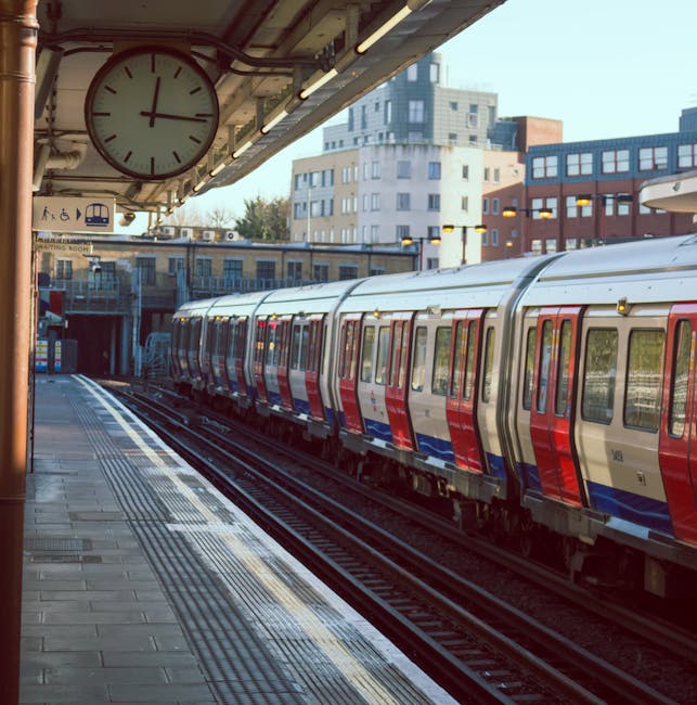 A view of a London Underground train with a silver body and red and blue accents, stopped at Oval station platform in Kennington. The platform has a tiled surface with tactile paving near the edge for safety, and a large round clock displaying the time as approximately 4:15. Overhead lighting illuminates the platform, with some visible wiring and structural components. In the background, urban buildings with modern architecture can be seen, and a section of the train tracks extends into a tunnel entrance. The scene is captured during daylight hours, with natural light reflecting off the train's exterior, supporting logistics related to moves and furniture transport at the station. As part of house removals or relocation services, visible elements suggest planning for the loading or unloading of goods, with the focus on the station environment and train transport infrastructure.