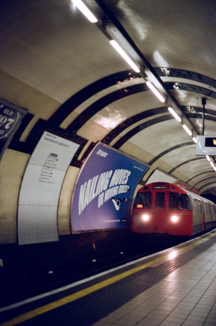 Inside an underground station tunnel with curved concrete walls and overhead lighting fixtures, a red London Underground train with its headlights on is approaching or arriving at the platform. Adjacent to the train, a large blue advertisement display promotes some form of service or event, partially visible behind the train. The platform's edge features a yellow safety line, and the station environment appears clean and well-maintained, indicating a busy urban transit setting. This image captures the typical scene of a London tube station, relevant to travel and transportation in house removals or moving logistics services related to metro access and timing, as seen in the page titled 'Oval station moves and removals timing and van access' on manwithvankennington.org.uk.