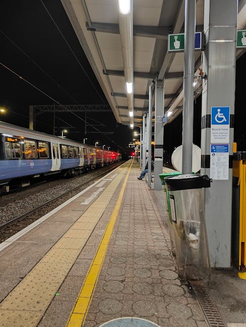 A view of a London Underground train with a silver body and red and blue accents, stopped at Oval station platform in Kennington. The platform has a tiled surface with tactile paving near the edge for safety, and a large round clock displaying the time as approximately 4:15. Overhead lighting illuminates the platform, with some visible wiring and structural components. In the background, urban buildings with modern architecture can be seen, and a section of the train tracks extends into a tunnel entrance. The scene is captured during daylight hours, with natural light reflecting off the train's exterior, supporting logistics related to moves and furniture transport at the station. As part of house removals or relocation services, visible elements suggest planning for the loading or unloading of goods, with the focus on the station environment and train transport infrastructure.