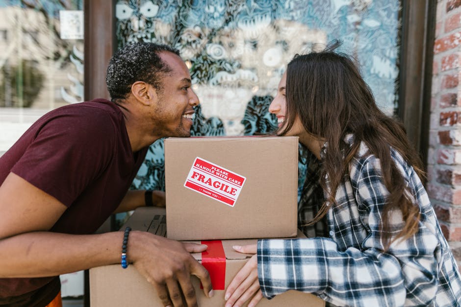 Two movers from Man with Van Kennington are engaged in a home relocation process outside a residential property. The man on the left, with short curly hair and wearing a dark red t-shirt, is smiling as he hands over a cardboard box labeled 'Fragile' with red and white warning tape, indicating careful handling. The woman on the right, with long brown hair and dressed in a blue and white plaid shirt, is also smiling and helping to support or receive the boxes. Both are positioned close to the doorway, surrounded by additional moving boxes, some wrapped in plastic, and a large moving van parked nearby, partially visible in the background. The scene is set outdoors, with a brick wall and window with patterned curtains behind them, suggesting they are in the process of packing or unpacking as part of a house move, with equipment such as trolleys and packing materials possibly nearby, typical of a professional removals and furniture transport service.